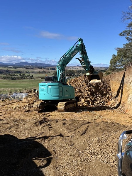 An excavator on a dirt block with rolling Tasmanian paddocks behind